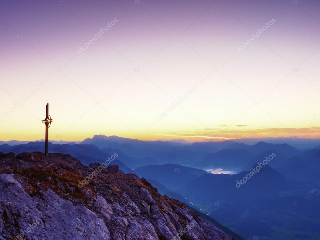 Steel crucifix at mountain peak in Alps. Sharp rocky summit, daybreak Sun in sky. 
