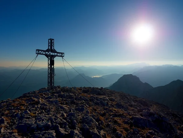 Steel crucifix at mountain peak in Alps. Sharp rocky summit, daybreak Sun in sky. — Stock Photo, Image
