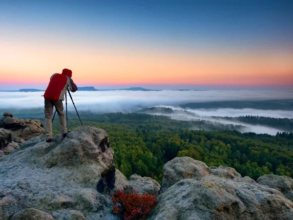 Fotógrafo tira fotos com câmera de espelho e tripé no pico da rocha. Queda paisagem nebulosa, primavera laranja rosa nebuloso nascer do sol no vale abaixo . — Fotografia de Stock