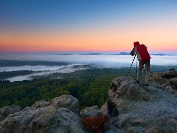 Fotógrafo tira fotos com câmera de espelho e tripé no pico da rocha. Queda paisagem nebulosa, primavera laranja rosa nebuloso nascer do sol em belo vale abaixo . — Fotografia de Stock