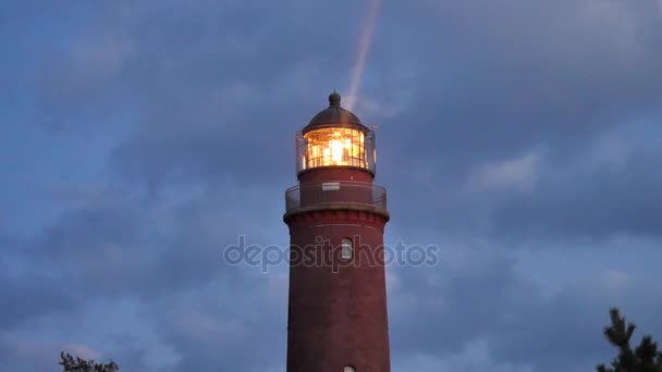 Farol histórico. Farol brilhante, dunas e pinheiros. Torre iluminada com forte luz de advertência, céu escuro no fundo. Torre do farol construída a partir de tijolos vermelhos . — Vídeo de Stock