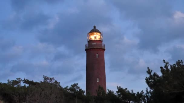 日没前に松の森の上の古い灯台をシャイニングします。強い警告灯で照らされたタワー。赤レンガからスポット ライトのガラスのカバーの周りの鉄の手すりが付いているギャラリーに建てられた灯台. — ストック動画
