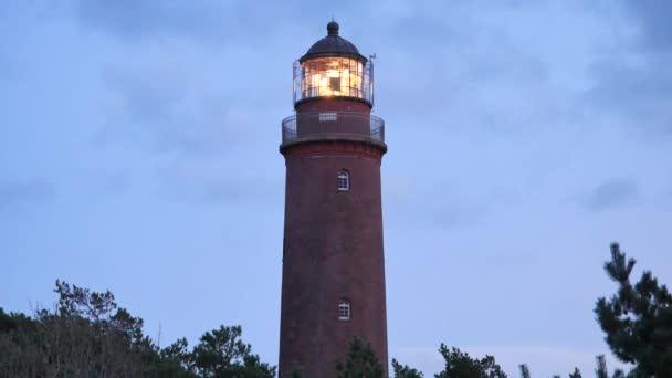 Leuchtender alter Leuchtturm über dem Kiefernwald vor Sonnenuntergang. Turm mit starkem Warnlicht beleuchtet. Leuchtturm aus roten Ziegeln, Galerie mit eisernem Geländer um Glasabdeckung des Scheinwerfers. — Stockvideo