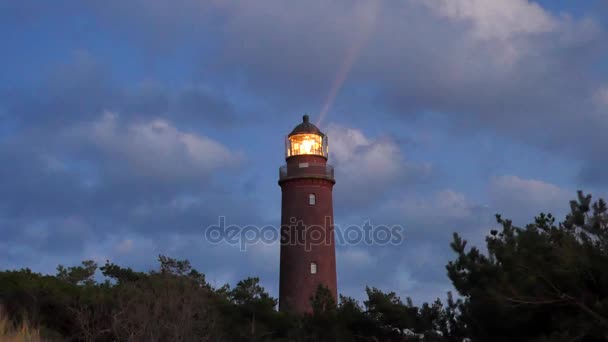 Shinning old lighthouse above pine forest before sunset. Tower illuminated with strong warning light. Lighthouse built from red bricks, gallery with iron handrail around glass cover of spotlight. — Stock Video