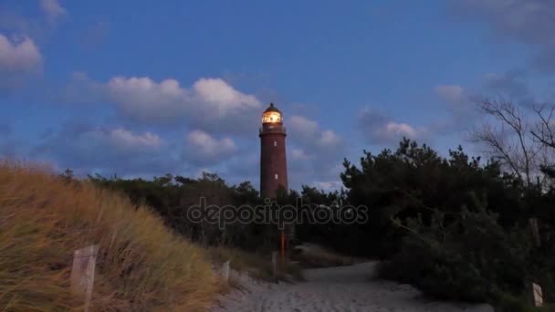 Shinning gamla fyren med mörka moln efter solnedgången i bakgrunden. Tower belysta med stark varningslampa. Fyren byggdes från rött tegel. Torr vass på sandstranden vandringsled med fot spår. — Stockvideo