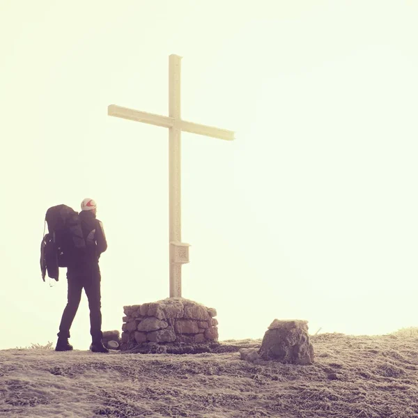 Tourist in heavy fog at cross memorial on mountain peak. Man is watching into misty Alpine valley bellow. Metal cross at a mountain peak. — Stock Photo, Image