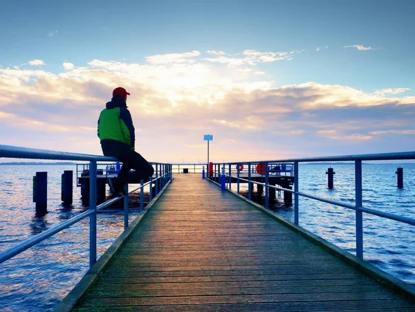 Mann in warmer Jacke und Baseballmütze sitzt auf Pier und genießt ruhige morgendliche See. Touristen entspannen. — Stockfoto