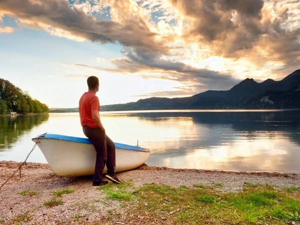 Tiired man in red shirt sit on old fishing paddle boat at mountains lake coast. — Stock Photo, Image