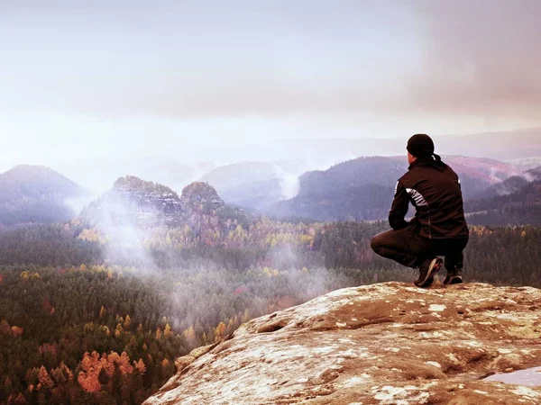 Turista em preto sentar-se na borda do penhasco olhando para enevoado vale montanhoso — Fotografia de Stock