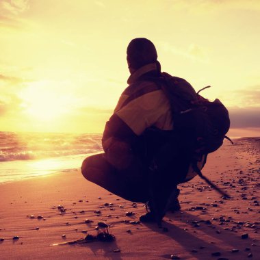 Man sit at evening sea. Hiker with backpack sit in squatting possition  along beach. Vintage Style Toned effect clipart