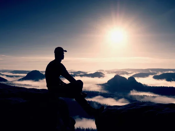 Silhouette of alone hiker in black and cap sit on cliff. Blue filter photo. Man looking to mis