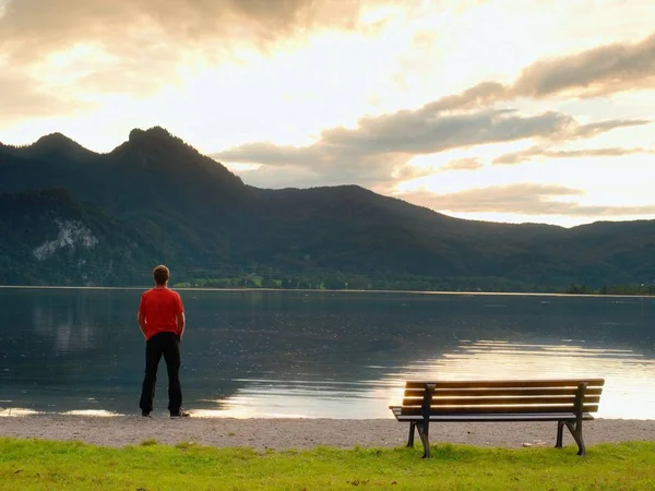 Tall man in red t-shirt at wooden bench at mountains lake coast. Dark clouds