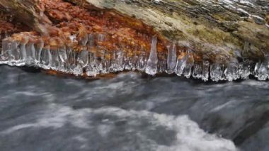Uzun buz sarkıtları dağ nehir karanlık soğuk suyun üstünde dur. Buz sarkıtları glitter akışı sütlü su üstünde. Düşmüş gövdeleri dallarında toz kar küçük gevreği tarafından karşılanmaktadır. 4k 3840 X 2160  