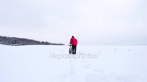 下に雪にマウンテン バイクを持つ男。バイクに乗る人は深い雪の中で自転車を押しています。穏やかな風と空気で小さな雪の結晶冬の曇りの日. — ストック動画