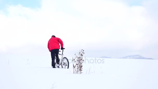 Man iker pousse le vélo dans la neige profonde au buisson sec de chardon. Journée d'hiver nuageuse avec un vent doux et de petits flocons de neige dans l'air . — Video