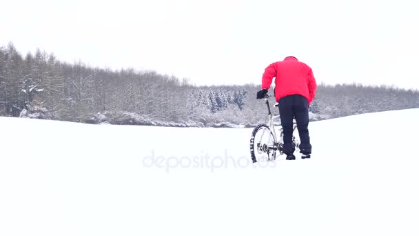 Man met mountainbike op besneeuwde geplaatst. Biker duwt fiets in diepe sneeuw. Bewolkt winterdag met zachte wind en kleine sneeuwvlokken in de lucht. — Stockvideo