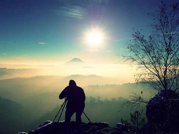 Manhã de inverno ensolarada. Fotógrafo preparando câmera no tripé. Rochas nevadas, no vale abaixo floresta folhas coloridas. Vista sobre neblina e nevoeiro vale para o sol . — Fotografia de Stock