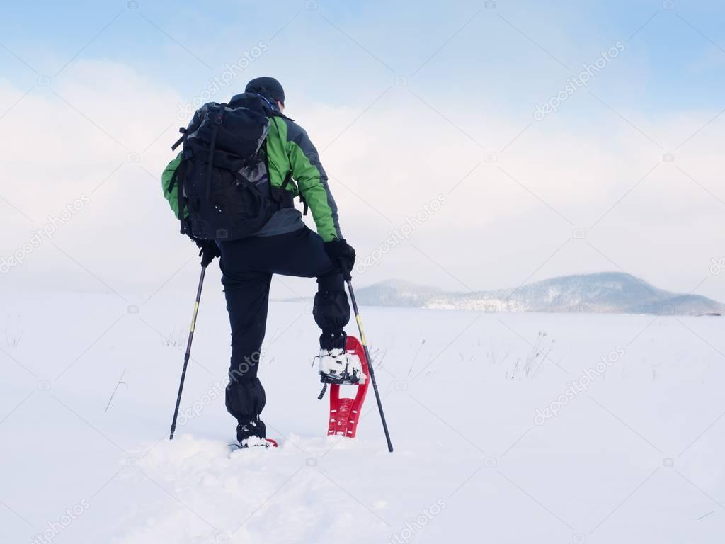 Man cleaning snowshoe. Hiker in green gray winter jacket and black trekking trousers walk in snowy filed. Snowshoeing in powder snow. Cloudy winter day, gentle wind brings small snow flakes 