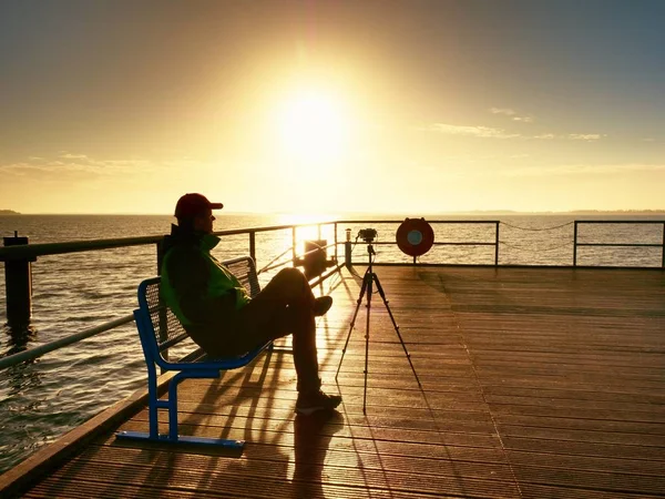 Hobbyfotograf sitzt auf Bank vor Kamera auf Stativ. Holzbrettkai — Stockfoto