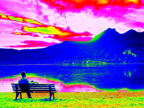 Infrared  photo. Alone man sits leg over on bench at mountain lake. — Stock Photo, Image