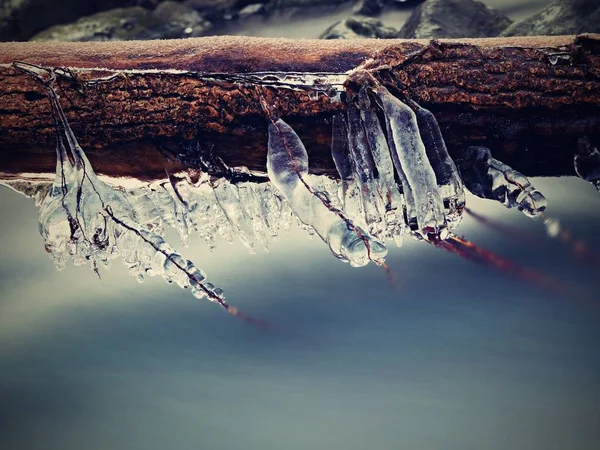 Largos carámbanos cuelgan sobre el agua helada oscura del arroyo de montaña . — Foto de Stock