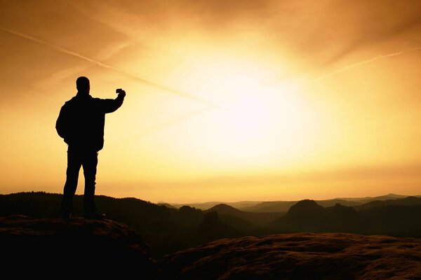 Man in mountains for a walk in evening with trekking poles.Sportsman in black with hands in pockets stand on the peak in rock empires park and watching over the misty and foggy morning valley to Sun. Silhouette of man.