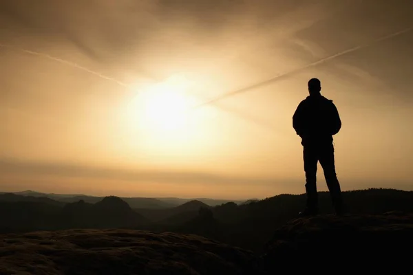 Deportista en negro con las manos en los bolsillos de pie en la cima en el parque de imperios de roca y mirando sobre el valle de la mañana brumoso y nebuloso al sol. Silueta del hombre . — Foto de Stock