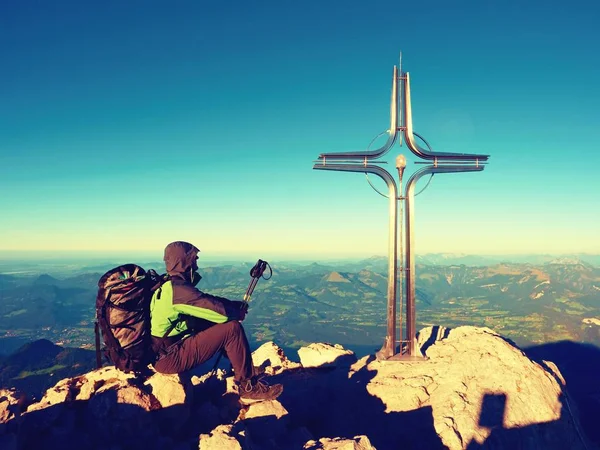 Tired hiker sit bellow crucifix on mountain peak. Iron cross at Alps mountain top