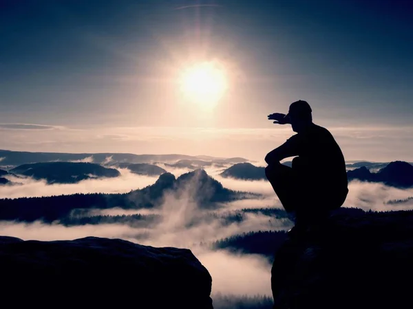 Silhouette of alone hiker in black and cap sit on cliff. Blue filter photo. Man looking to mis — Stock Photo, Image