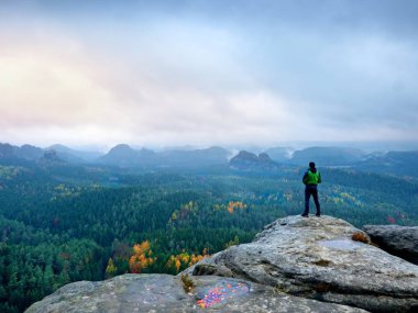 Uzun yürüyüşe çıkan kimse yeşil windcheater, kap ve karanlık trekking pantolon dağ en yüksek kayanın üzerinde durmak.