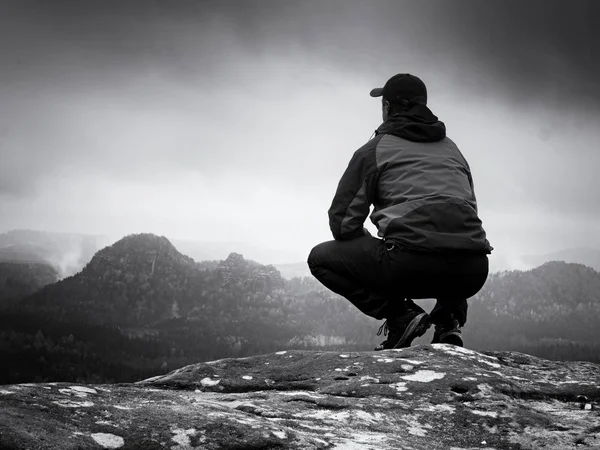 Νεαρός άνδρας με τα μαύρα κάθονται στην άκρη του βράχου, και ψάχνει να ομιχλώδη κοιλάδα — Φωτογραφία Αρχείου
