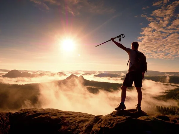 Alto zaino in spalla con pali in mano. Sunny daybreak nebbioso in montagne rocciose. Escursionista con zaino in piedi sul punto di vista roccioso sopra valle. Vivido e forte effetto vignettante . — Foto Stock