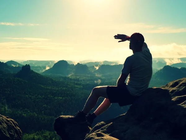 Człowiek siedzieć na szczycie skały. Turysta przesłanianie oczu z podniesione ramię, oglądając kolorowe mgła i mgła w dolinie lasu. — Zdjęcie stockowe