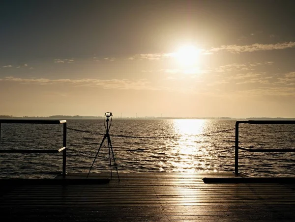 Tripé com câmera pronta no cais, Sol acima do oceano. Placa de toupeira de madeira vazia — Fotografia de Stock