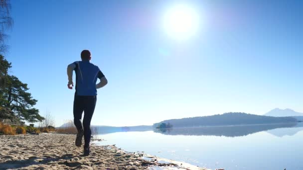 Hombre alto con gafas de sol, gorra de béisbol roja y ropa deportiva negra azul está corriendo y haciendo ejercicio en la playa al mediodía. Deporte hombre activo estirar las piernas y el cuerpo en la playa del lago de montaña . — Vídeos de Stock