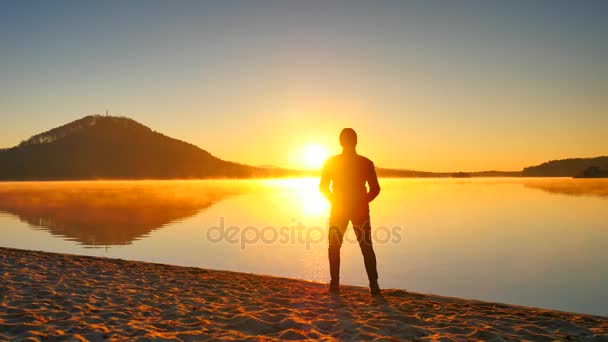 Homem alto com óculos escuros e boné escuro está correndo na praia ao pôr do sol. Silhueta de esporte homem ativo correndo e exercendo na praia do lago de montanha. Laranja pôr-do-sol céu fundo . — Vídeo de Stock