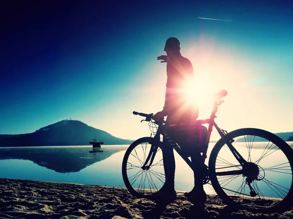 Sylwetka sportowca gospodarstwa rowerów na plaży nad jeziorem, kolorowy zachód słońca niebo pochmurne, w tle — Zdjęcie stockowe