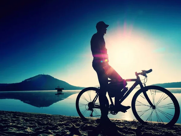 Sylwetka sportowca gospodarstwa rowerów na plaży nad jeziorem, kolorowy zachód słońca niebo pochmurne, w tle — Zdjęcie stockowe