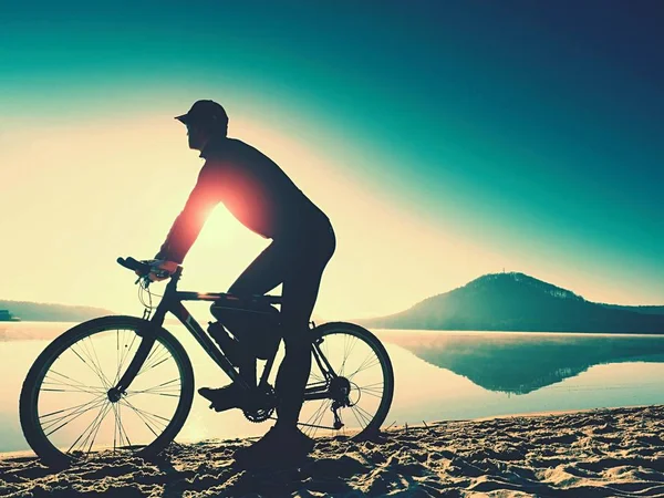 Silhueta de desportista segurando bicicleta na praia do lago, colorido céu nublado pôr do sol no fundo — Fotografia de Stock