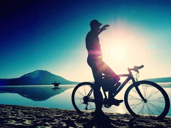 Silhueta de desportista segurando bicicleta na praia do lago, colorido céu nublado pôr do sol no fundo — Fotografia de Stock