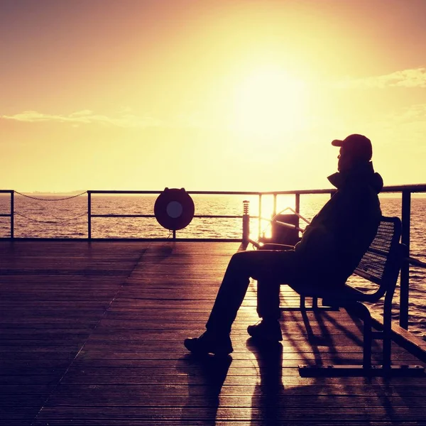 Mann in warmer Jacke und Baseballmütze sitzt auf Pier und genießt ruhige morgendliche See. Touristen entspannen. — Stockfoto