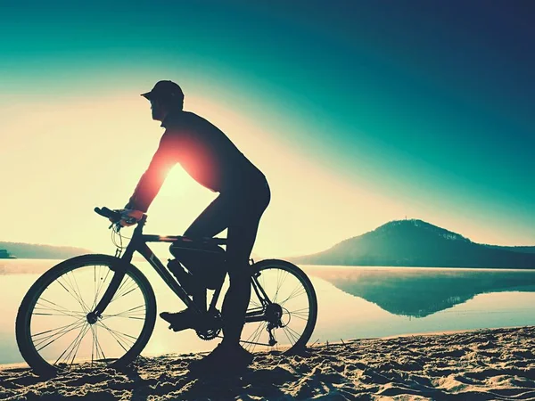 Silhueta de desportista segurando bicicleta na praia do lago, colorido céu nublado pôr do sol no fundo — Fotografia de Stock