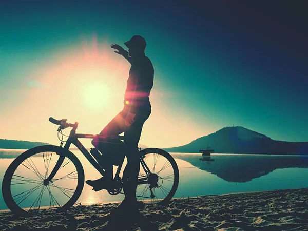 Silhueta de desportista segurando bicicleta na praia do lago, colorido céu nublado pôr do sol no fundo — Fotografia de Stock