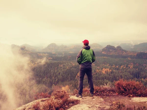 Человек турист остается на пике острой скалы. Один турист в красной кепке и зеленой куртке наслаждаться видом — стоковое фото
