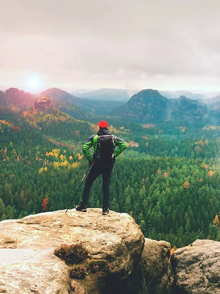 Guía turística en la cumbre con bastones en la mano y pesada mochila. Caminante verde jakcet nad gorra roja —  Fotos de Stock