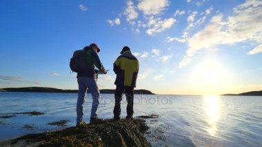 Balık tutma ve yürüyüş hakkında konuşuyorsun. Genç balık adam denizde uzun yürüyüşe çıkan kimse adam trekking Polonyalılar ve yeşil sportif sırt çantası ile konuşur. Güzel gün batımı ile balıkçılık zevk erkekler