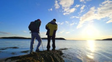 Balık tutma ve yürüyüş hakkında konuşuyorsun. Genç balık adam denizde uzun yürüyüşe çıkan kimse adam trekking Polonyalılar ve yeşil sportif sırt çantası ile konuşur. Güzel gün batımı ile balıkçılık zevk erkekler