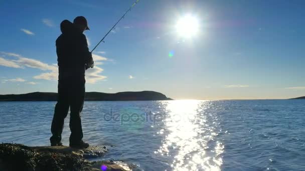 Un joven con barba en ropa negra al aire libre está pescando mientras está de pie en la costa rocosa. Agua con gas brillante — Vídeos de Stock