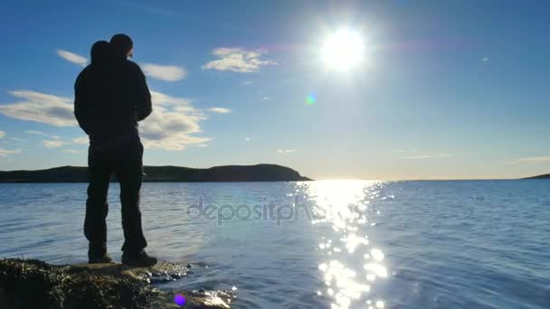 En ung man med skägg i svart utomhus kläder är att fånga fisk stående på steniga stranden. Ljusa kolsyrat vatten — Stockvideo
