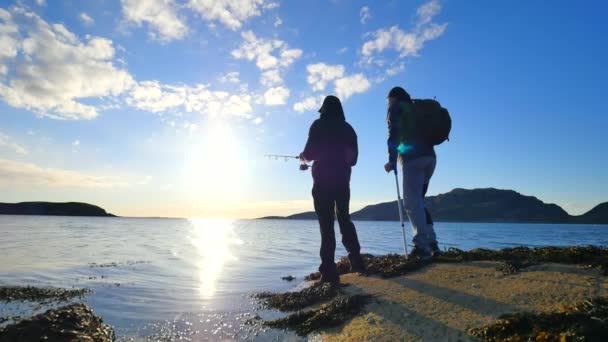 Los amigos hablan de pesca y salud. Joven pescador trabajador en piedra al nivel del mar. Caminante con muletas medicinales y pierna en frenos de rodilla funcionales. Hombres disfrutando de la pesca con hermosa puesta de sol — Vídeos de Stock